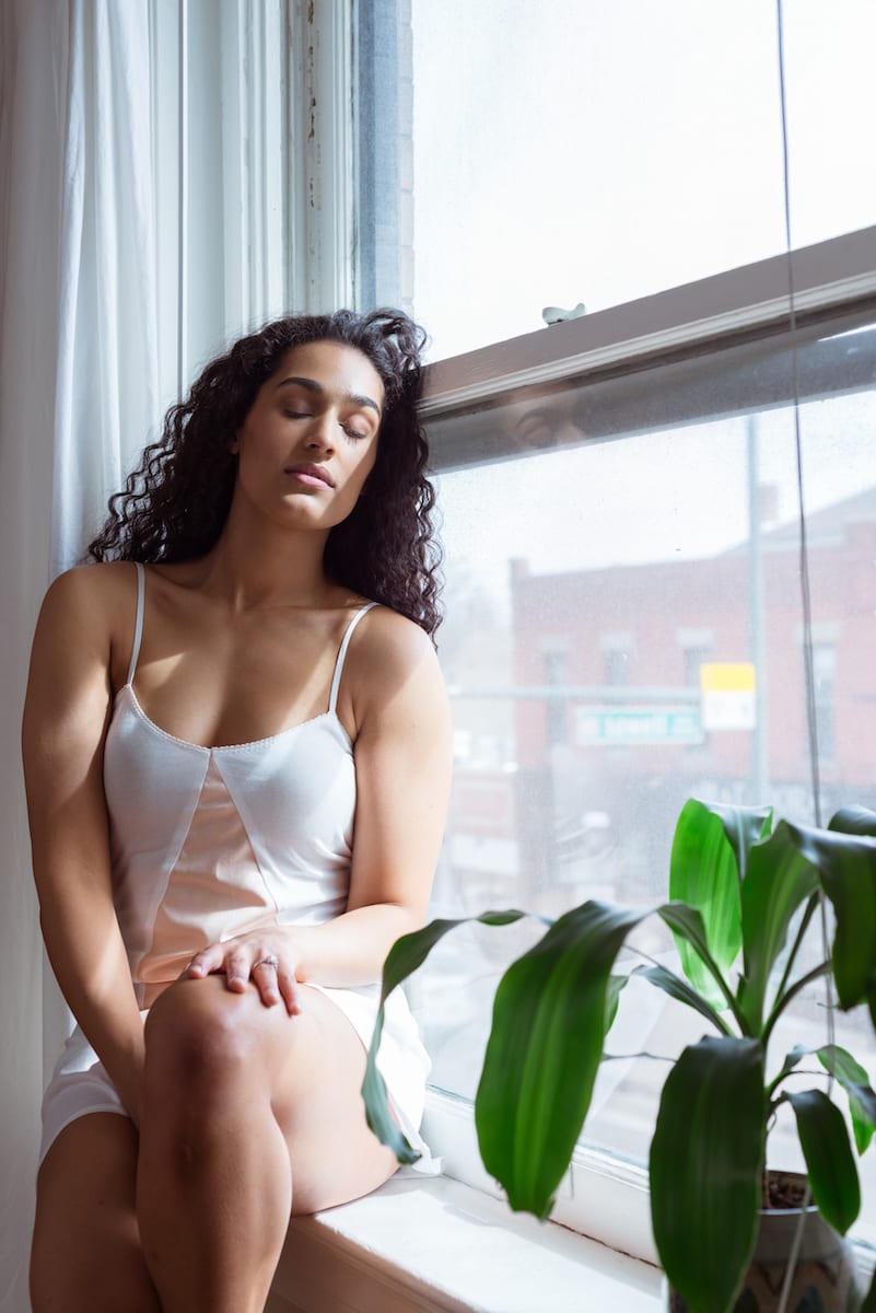 boudoir | Boudoir Photography | model | From the Hip Photo | Model in white and peach slip sits on window sill with eyes closed and head presses against the window