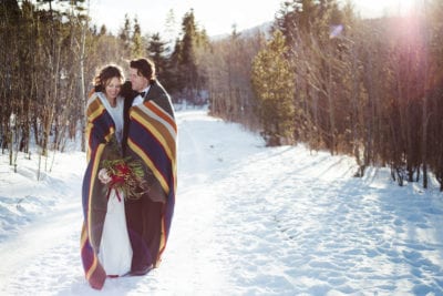 bride and groom walk through a snowy alley wrapped in a large blanket