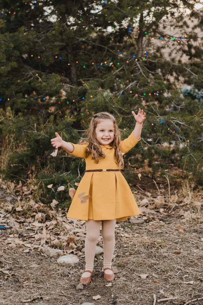 Little girl smiles and poses in front of Christmas tree outdoors