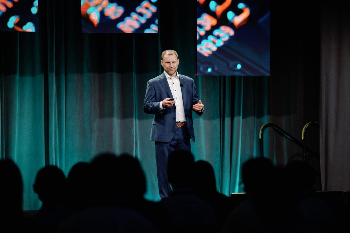 Keynote speaker in blue suit gesturing on stage with screens behind him