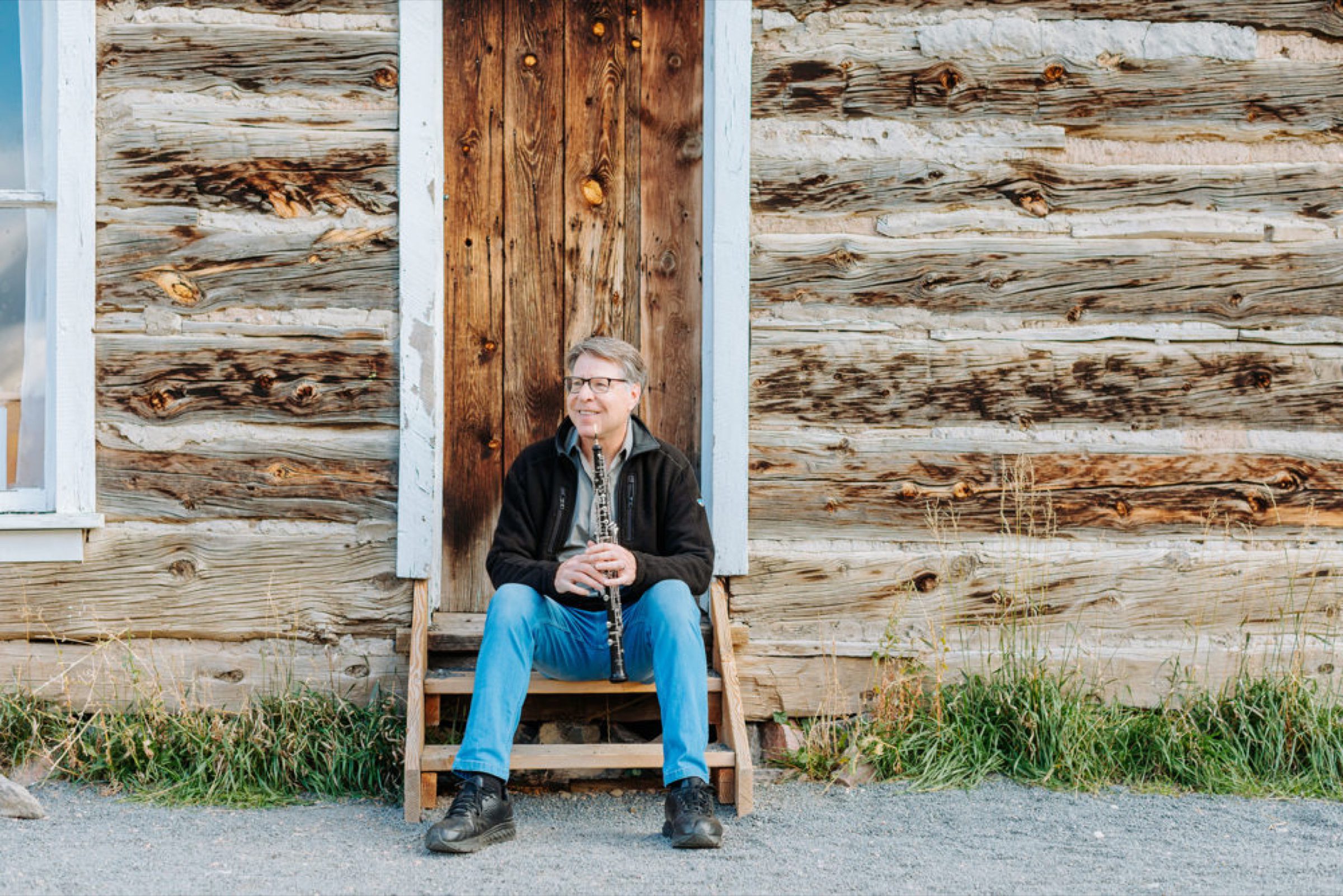 Oboist on the steps of a weathered log cabin in tall prairie grass