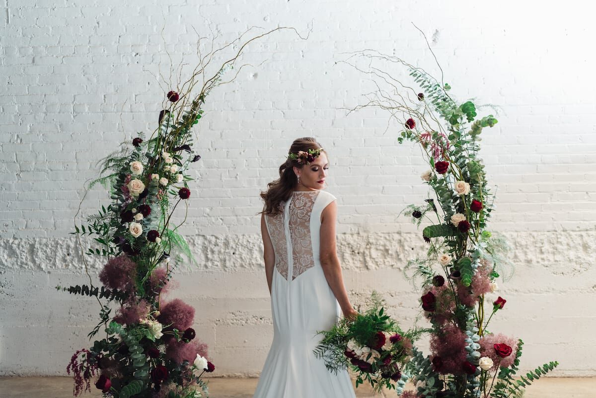 A bride beside a floral installation of dried grasses and deep red blooms against white brick at Moss Denver