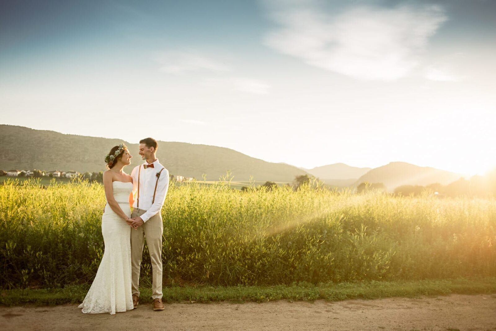 Bride and groom holding hands in tall summer grass at sunset, with the foothills behind them at Chatfield Farms